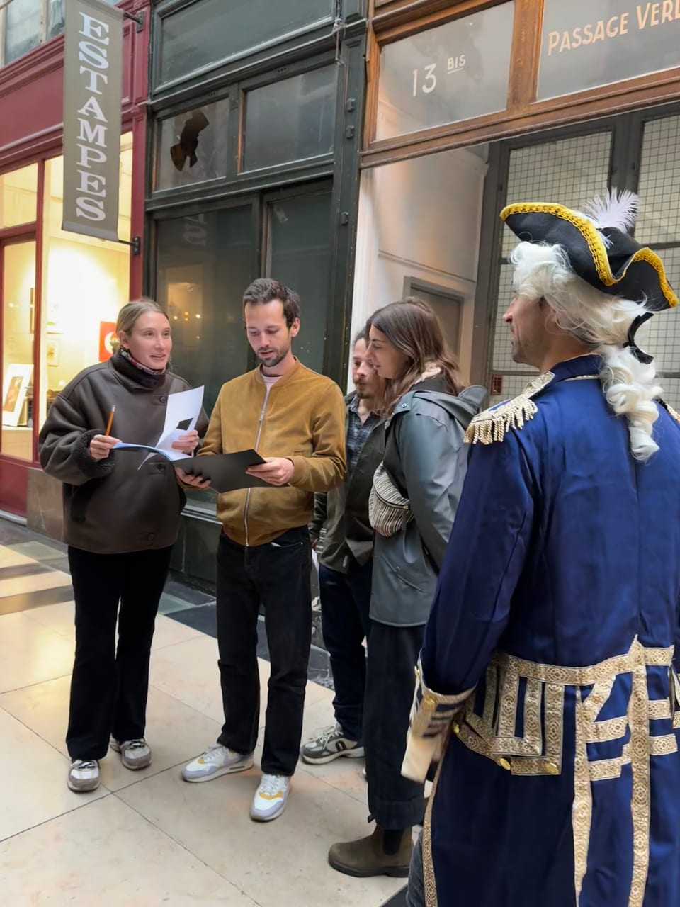 Charles Oudin en costume et un groupe de joueurs penchés sur une énigme, devant le passage Verdeau