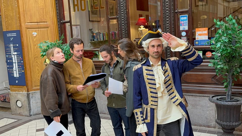 Le game master en costume au premier plan, un groupe en action derrière lui