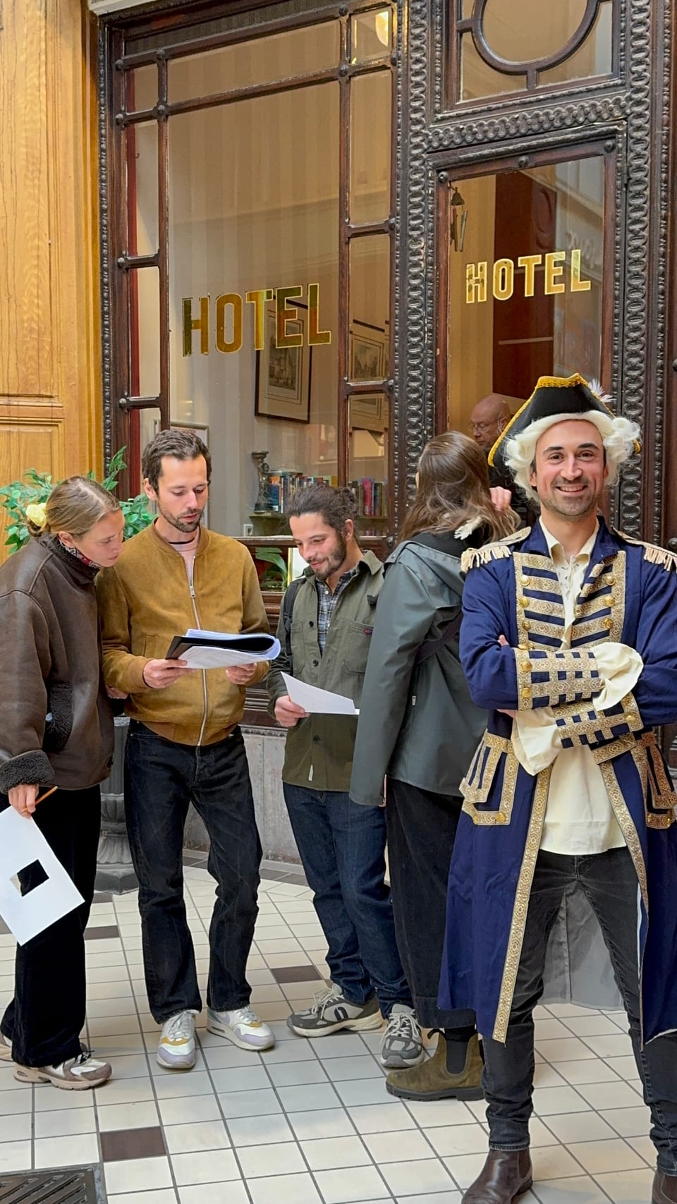 Charles Oudin en costume, bras croisés et souriant, devant un groupe d'amis penchés sur une énigme dans un passage couvert